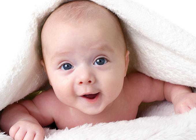 Baby Cute - This image captures a close-up view of a newborn baby. The baby's eyes are open, gazing unfocused, likely because the eyes are not fully developed at birth. The baby's hands rest near its face, giving the appearance of smiling. The baby is snuggled on its side and is draped with a white blanket, possibly posed for a portrait given the soft lighting and focus on the face. This image conveys a sense of innocence and vulnerability typically associated with newborns.