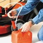 The image shows a person handling a lawn mower. The person is holding the mower upside down and appears to be engaged in some sort of maintenance or cleaning task. They are wearing a blue jacket, and there is an orange bottle connected to a tube which seems to be part of the lawn mower. The setting seems to be a domestic environment with a motorcycle parked in the background. The focus of the image is on the interaction between the person and the lawn mower.