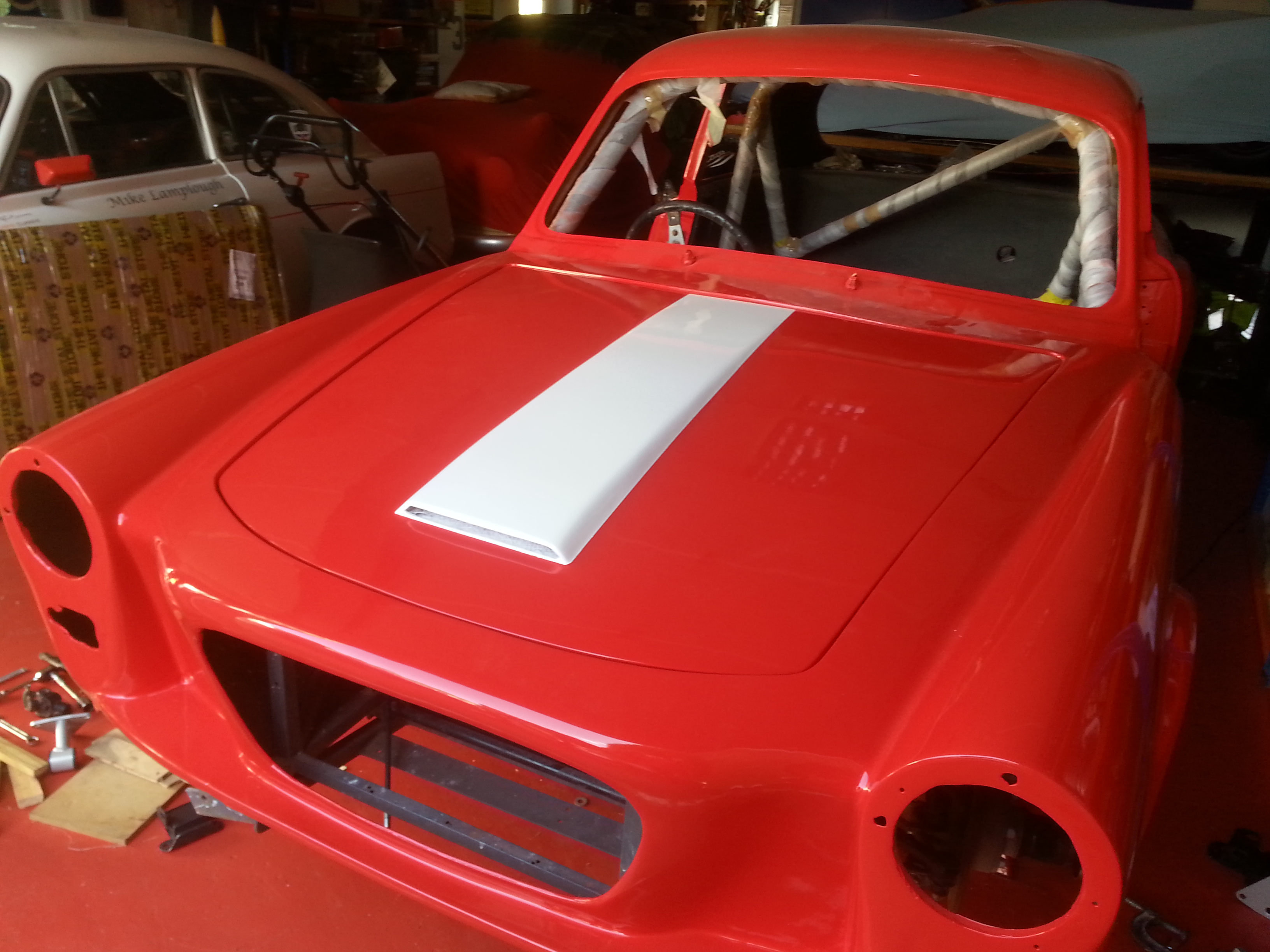 Pistonheads - The image shows a classic red race car that appears to be under restoration or renovation. It's situated indoors, likely within a garage or workshop space. The car features a prominent white stripe along its side, which is often associated with racing cars. The bodywork of the car looks freshly painted and possibly modified, as indicated by the missing hood and the open front panel that reveals engine components. There are also some tools visible in the background, suggesting a work-in-progress environment.