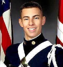 The image features a young man who appears to be a military servicemember. He is wearing a black and white military uniform with visible rank insignia and is holding and partially concealing a Stetson hat with ribbons. He is positioned in front of two American flags, which are at full mast behind him, suggesting a level of formality or solemnity in the photo.