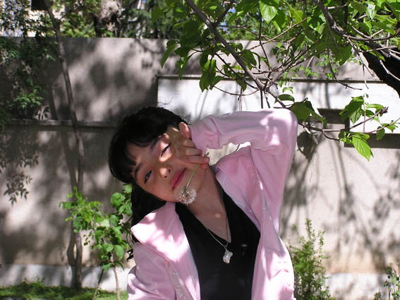 The image portrays a young woman outdoors, dressed in a light pink jacket, standing in a sunlit area. She appears to be in a moment of relaxation, holding what seems to be a white flower delicately to her nose, as if to smell its fragrance. She has her arm gently resting on a tree branch, suggesting a casual and peaceful setting. The background is filled with trees and a fence, providing a natural and possibly urban backdrop to the scene.