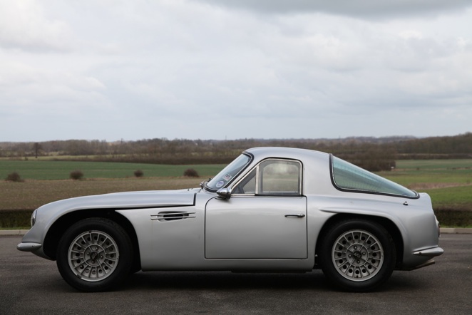 Early TVR Pictures - Page 35 - Classics - PistonHeads - The image showcases a classic Aston Martin DB5 sports car parked on a road. The car's sleek silver body gleams under the clouds, and the hood is emblazoned with the iconic DB5 logo. The Aston Martin DB5 is positioned on the side of the road, facing to the right, with its top-down. The backdrop is a serene landscape featuring green fields and distant hills under a partly cloudy sky.