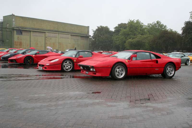 Speciale's big brother has arrived - Page 6 - Ferrari V12 - PistonHeads - The image shows a line of red sports cars parked in a parking lot. The cars are similar in style and color, suggesting they may be a part of the same collection or event. They are parked close together, some also touching, which could indicate they were not arranged intentionally but rather randomly. In the background, there are some taller structures that could be buildings, providing an urban setting for the scene. The ground appears to be wet, possibly due to recent rain, which reflects on the cars' surface and adds visible wet spots to the image.