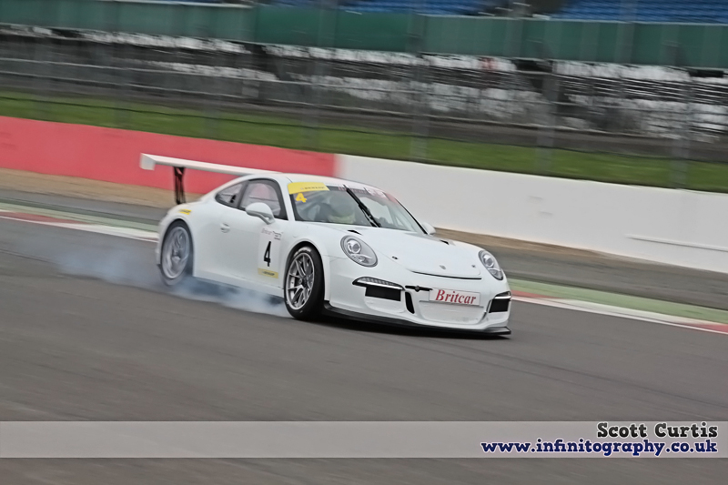 Silverstone Pics - Page 1 - Britcar - PistonHeads - The image captures a dynamic scene of a white Porsche 911 rally car in action on a racetrack. The car is skillfully navigating a right turn, its rear wheel lifted slightly off the ground as it leans into the curve. The racetrack is lined with a red and white barrier, adding a splash of color to the scene. In the background, a crowd of onlookers can be seen, reinforcing the intensity of the event. Additionally, there's text in the image that says "INFINITOGRAPHY.co.uk SCOTT CURTIS" which could be related to the photographer, indicated sponsorship, or belong to a website associated with the event.