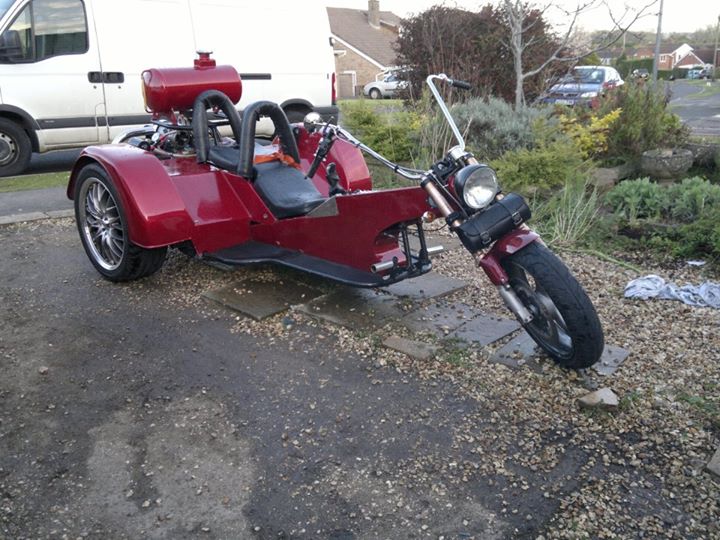 Pistonheads - This image features a striking red three-wheeled motorcycle parked on a gravel driveway. The vehicle stands out against the backdrop of a parking lot filled with parked cars, including a van immediately to the left of the motorcycle. The motorcycle sports a horn on the front, and its design includes a sidecar attachment on the left side, hinting at potential passenger transportation. The scene gives off a quiet, momentary stillness, with no people or significant action visible. The focus is solely on the vibrant, three-wheeled motorcycle, making it the central subject of this image.