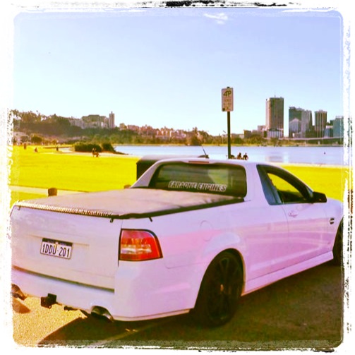 Ute Numbers in the UK??  - Page 1 - HSV & Monaro - PistonHeads - The image depicts a white truck parked on a road near the coastline. The area features a grassy field with a few people scattered about, seemingly enjoying their time. The cityscape in the background is adorned with trees lining the coast, and a bridge can be seen across the water. The truck bears a personalized slogan on its body, which appears to be a mix of text and graphics, although the specific content is not legible. The atmosphere suggests a leisurely day out, with the sea visible in the background.