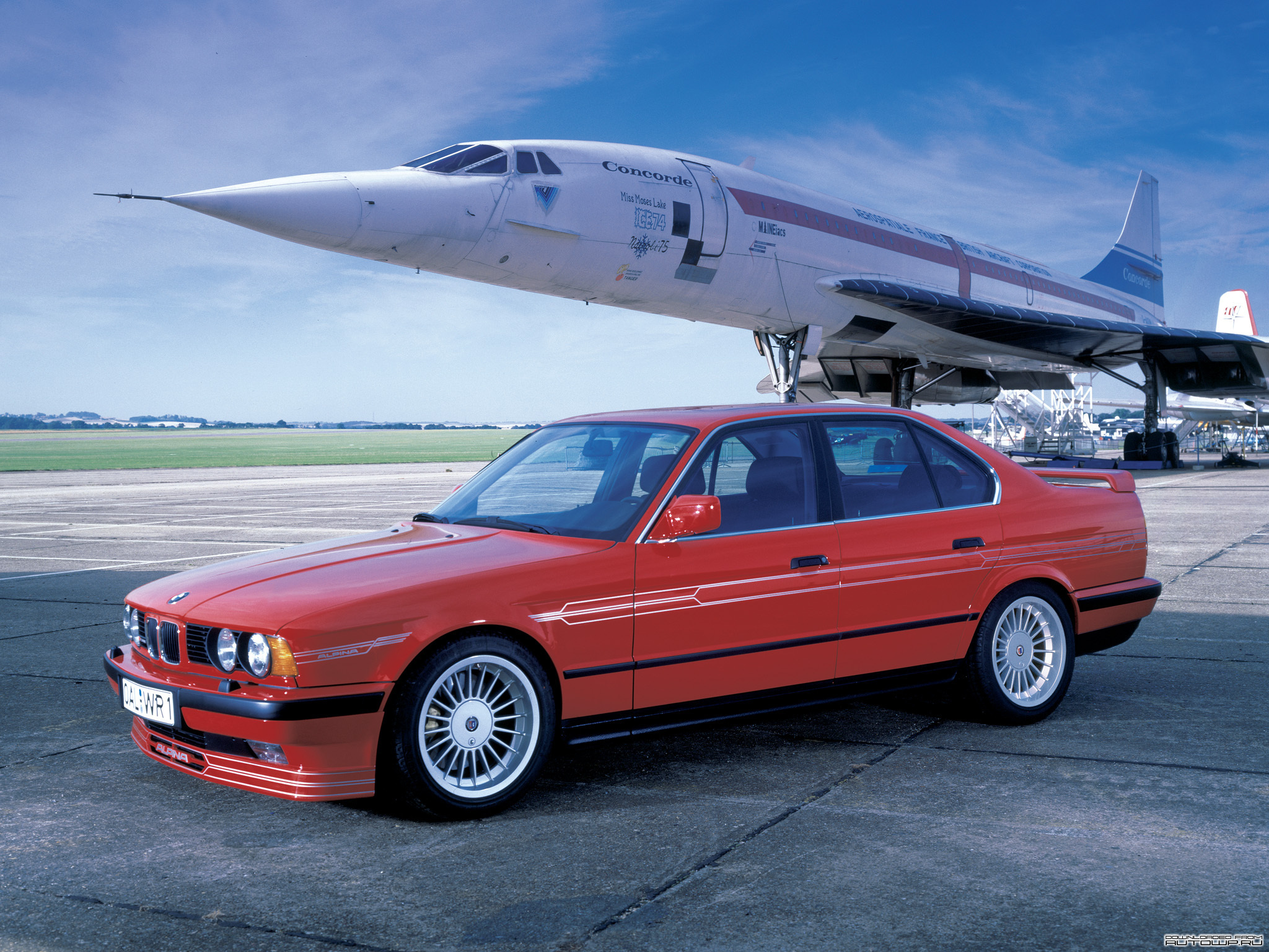 RE: Spotted: 1990 Alpina B12 750i - Page 5 - General Gassing - PistonHeads - The image features a vibrant red BMW car parked on an airport tarmac. Looking beyond the car brings us to a Concorde SST, the car of the future, available at a local dealership. The car and plane are situated on a runway, evoking a sense of travel and adventure. The BMW car is facing the viewer, showcasing its sleek design and polished exterior, while the Concorde is facing left, as if ready to take off for a journey through the skies.