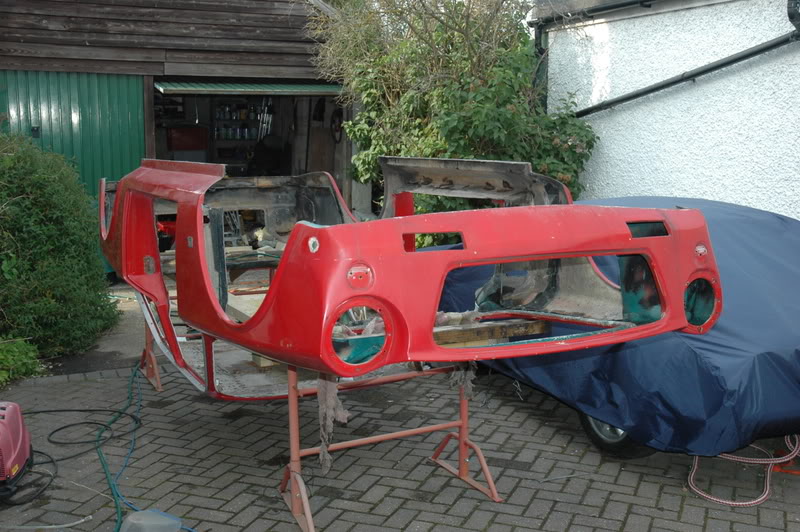 Amazon, P6 or left field choice? - Page 3 - Classic Cars and Yesterday's Heroes - PistonHeads - The image shows a red car that appears to be in the process of being painted or restored. The car is missing its bodywork, revealing a metal frame, and it resides on a paved lot with grey stone bricks in the foreground. In the background, a white building can be seen along with a partially covered car atop a blue tarp. There's a sense of workmanship and craftsmanship, suggesting a potential custom or classic car restoration project.