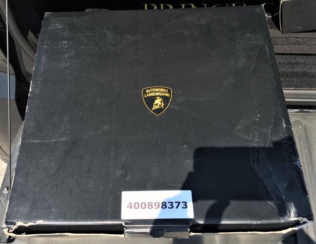 2013 Gallardo LV63 LXH / X8 DRG / 4 TBD / M5 KPP / M477 PET? - Page 1 - Gallardo/Huracan - PistonHeads UK - The image shows a black box with a distinctive logo, resembling a shield and a lion, placed on the trunk of a vehicle. This logo suggests it is a product from the brand Lamborghini. In front of the box, there is an object that looks like a shovel or possibly some kind of tool. The box itself appears to be open or damaged, with part of its side broken off, revealing the interior. At the bottom right corner of the image, there's a white tag with black text and numbers, which might indicate a purchase or shipping label. The background is blurred, but it seems to be an outdoor setting with daylight illuminating the scene.