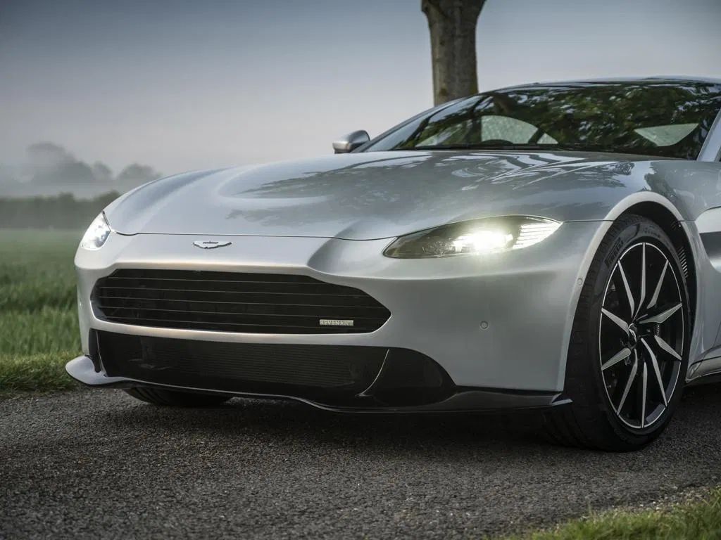 Pistonheads - The image features a sleek, silver-colored Aston Martin sports car positioned on what appears to be a wet road at night. The vehicle is parked in the middle of the frame, facing towards the left side of the photo, and it's captured from an angle that showcases its front and side profile. The car's headlights are on, illuminating part of the road ahead. The background is dark, which contrasts with the car, emphasizing its presence in the image. There's a subtle reflection on the wet road surface, suggesting recent rainfall.