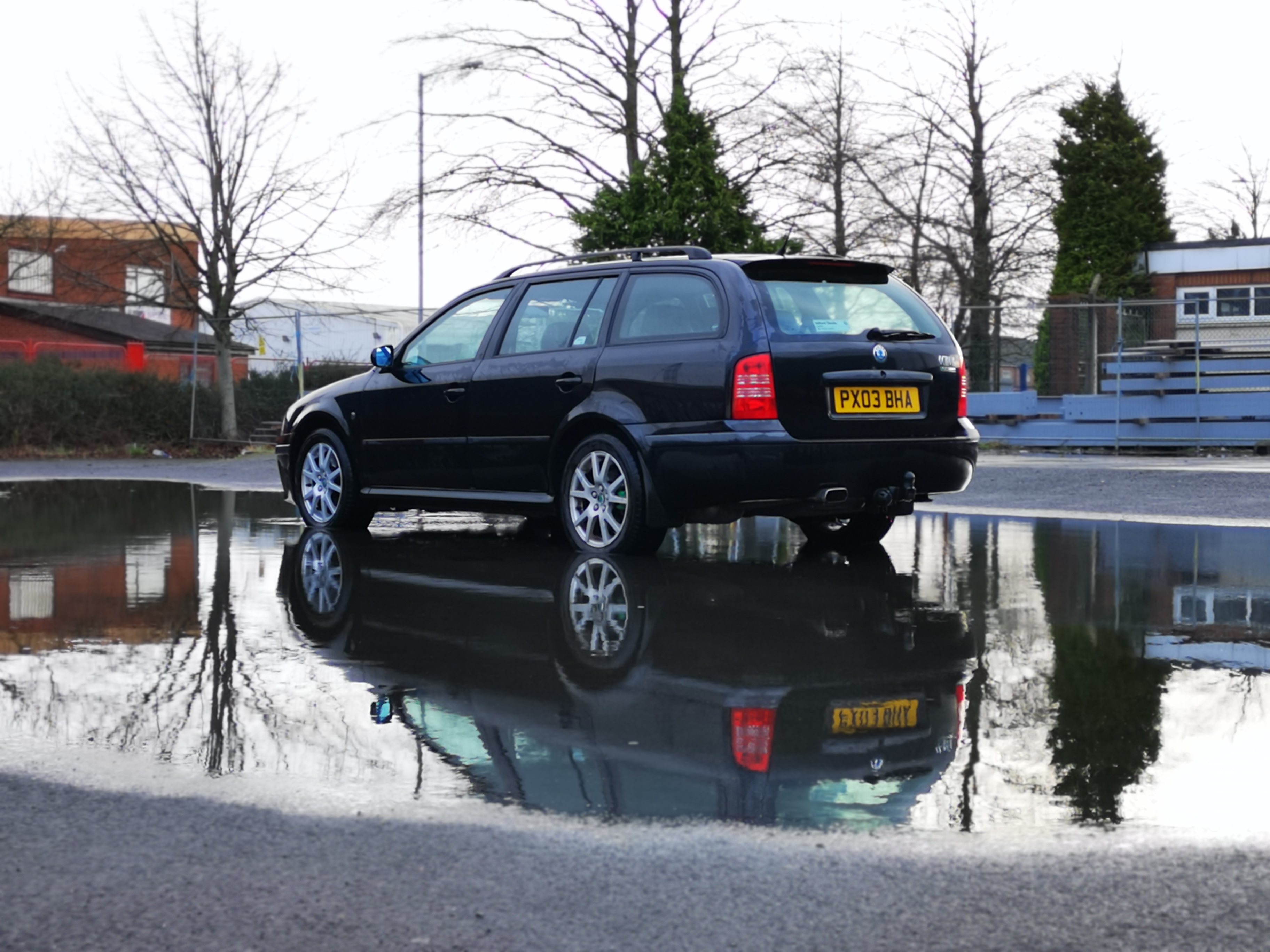 RE: The best fast estates to buy in 2020 - Page 13 - General Gassing - PistonHeads - The image displays a car parked on a wet road with a puddle in front of it. The car appears to be a dark-colored station wagon, possibly from the late 20th century based on its design. The surrounding environment suggests an urban setting, with buildings and trees visible behind the vehicle. There's a reflection of the car in the water, adding a symmetrical element to the scene. A set of keys is also seen resting on the hood of the car, indicating someone might be nearby.