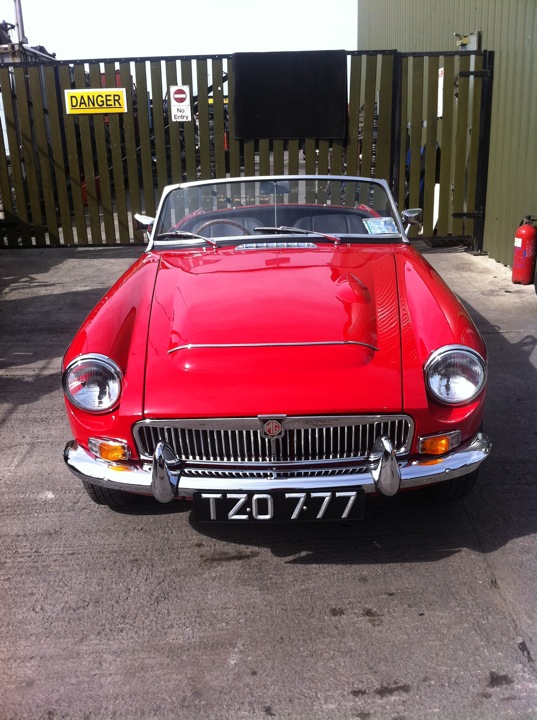 1969 Irish MG-C Roadster - Page 1 - Classic Cars and Yesterday's Heroes - PistonHeads - The image features a classic, two-door, convertible sports car painted in a bright, glossy red. It's parked in a lot, facing directly towards the camera. The car's front grille is visible, adorned with a silver emblem resembling a shield or circle. In the background, we can see a green fence and a gray building, along with a yellow sign with red text indicating some form of warning or danger, which includes an outlined white shape resembling a child for some reason. The license plate of the car displays the number '777', visible on the front.