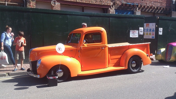 Camberley Town Centre car show - Page 1 - Thames Valley & Surrey - PistonHeads - The image captures a lively street scene featuring a vibrant orange vintage truck as the main subject. The truck, which appears to be a restored classic model, is parked on the side of the street. It is an eye-catching pop of color against the more neutral tones of the surroundings. There are three people in the image, two standing next to the truck, and one possibly engaged in a store purchase in the background. The street scene and the presence of the vintage truck suggest a possible public event or a place where vintage cars and trucks are showcased.