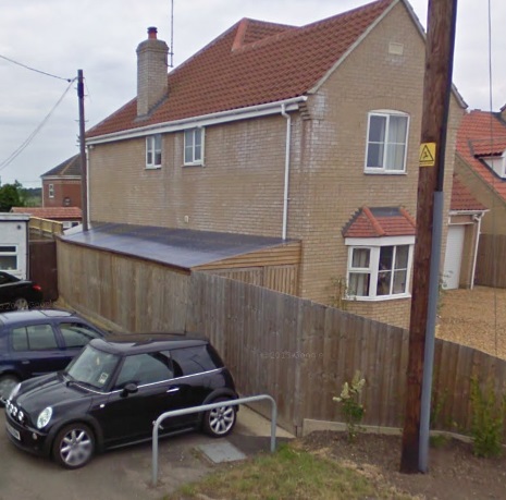 Another boundary dispute  - Page 1 - Speed, Plod & the Law - PistonHeads - The image shows a small black Mini Cooper parked in a residential area. The car is positioned near the sidewalk on the passenger side. The vehicle is close to a fence and appears to be a two-door model. Behind the car, there is a two-story brick building with a sloping roof, windows, and a small porch area. To the right, there's a utility pole with some markings on it. The sky is overcast, suggesting cloudy weather.