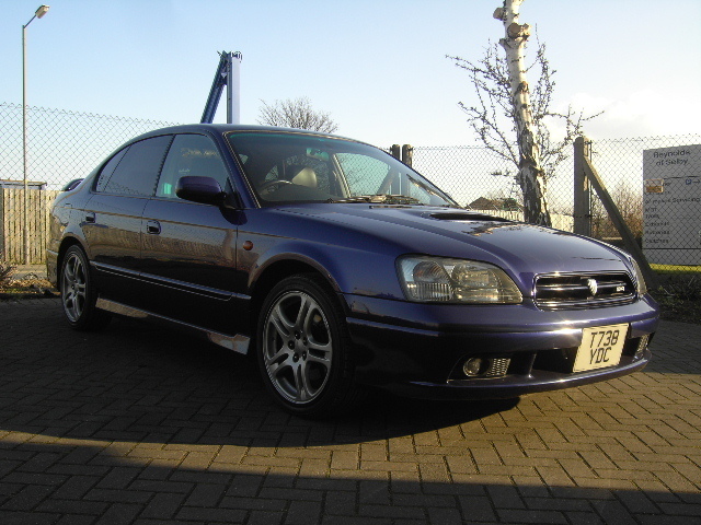 Pistonheads - The image shows a dark blue car parked on a bricked area with a fence in the background. The car appears to be a model from the late 20th century, possibly a sports sedan due to its sleek design and large grille. There are signs posted near the fence, including one with a company logo. The sky is clear, suggesting a sunny day. The car is turned slightly to the side, allowing a view of its profile.