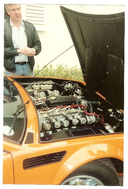 Bob Wallace 1938 - 2013 - Page 1 - Lamborghini Classics - PistonHeads - In the image, a man stands next to an opened light orange sports car. Its hood is up, revealing a hot rod engine with several throttle bodies dominating the center. The man, dressed in a casual black jacket, appears to be checking on the car or perhaps admiring the intricate engine work. The setting is outdoors, with some greenery visible in the background, suggesting a garden or a park might be nearby. The relative position of the man and the car indicates he is closely inspecting the vehicle's mechanical details.