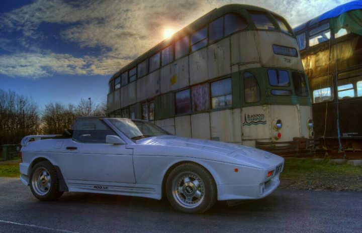 Random TVR pics - Page 305 - General TVR Stuff & Gossip - PistonHeads - The image features a striking scene on a highway. Dominating the left side of the image is a silver sports car, its sleek design and shiny finish reflecting the clear blue sky above it. Behind the sports car, a white and green double-decker bus stands tall, seemingly in tow. The background is characterized by a roadside lot, gathered under a few scattered trees, adding a touch of nature to this urban setting. A small building can be seen further down the road, adding another layer of intrigue to this picturesque scene. The image captures a moment of stillness, with both the sports car and the bus parked stationary, waiting for what lies ahead.