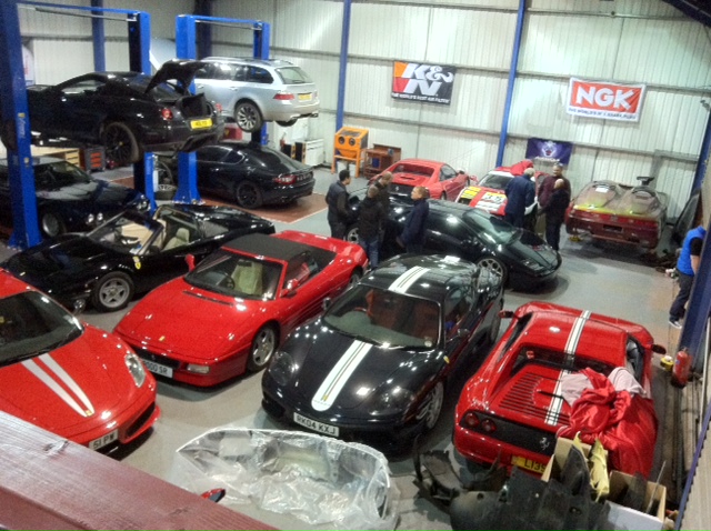 The GT Cafe Gallery - post your meeting photos here - Page 1 - North West - PistonHeads - This is a photograph of an indoor facility with a variety of sports cars on display. The cars are arranged randomly throughout the space, some indicating a history of racing, as suggested by the race numbers and sponsor logos on some, which include Pirelli and NK. There are at least ten cars in total, ranging in color from red to black, with some featuring distinctive stripes like a red and white Ferrari. People can be seen working within the area. The setting appears to be a garage or storage area, as indicated by the presence of equipment racks and large doors in the background. It's a bright day outside, as suggested by the quality of light filling the space.