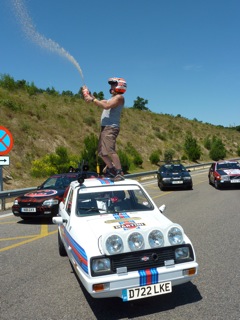 "Chip run cars win in Barcelona" - Page 1 - East Anglia - PistonHeads - This image captures a thrilling stunt performed by a man on top of a vehicle. The man is riding a car-like contraption, which is mounted on top of a sports car where he is standing. The contraption and the car it is attached to are adorned with a striking blue, white, and black color scheme. They are situated on a two-lane highway, with several other cars visibly present behind them. The backdrop of the scene is a clear blue sky, creating a stark contrast with the vehicle's vibrant colors. The action captured in the image is adrenaline-filled and demonstrates the man's daring spirit.