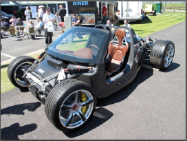 Carrera Pistonheads - The image shows a futuristic-looking vehicle parked on a paved surface. It's a sleek, black electric car with a large cockpit and spoked wheels. The car is on display at an outdoor exhibition, as evidenced by the tent in the background which says "Diner." There are a few people standing nearby, but they are only partially visible. The setting appears to be an outdoor area with grass around the car, indicating it's part of the exhibition premises.