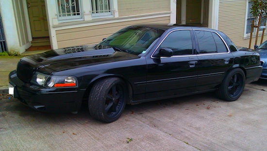 A car with a surfboard strapped to the back of it - Pistonheads - This image features a black sedan parked in front of a beige house with white trim. The car has a distinctive stance, lowered on its back wheels, and is equipped with a custom bumper and wheel rims, which gives it a distinctive look. It's a scene of a quiet, suburban neighborhood. The sky is overcast, suggesting it might be a cool day. The car is the main focus of the image, with its sleek lines and unique modifications.