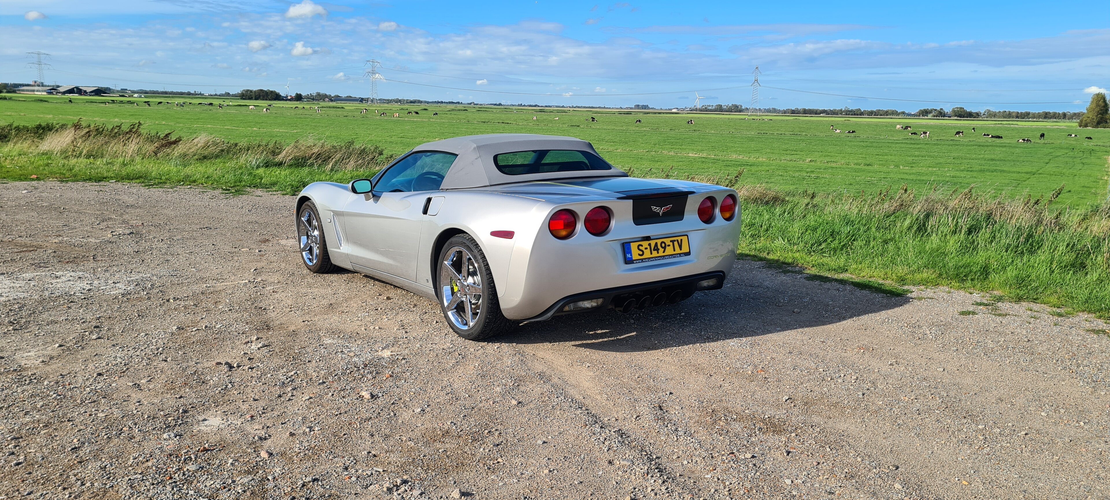 The £7700 Corvette C6 - Page 29 - Readers' Cars - PistonHeads UK - The image depicts a convertible sports car parked on what appears to be a gravel or dirt road. It is daytime with clear skies, and the vehicle has its top down, revealing a well-maintained interior. The car is sleek with a long hood, large alloy wheels, and a distinctive design that suggests speed and performance. The surrounding landscape includes open fields, indicating a rural or semi-rural setting. There's no visible text on the image.