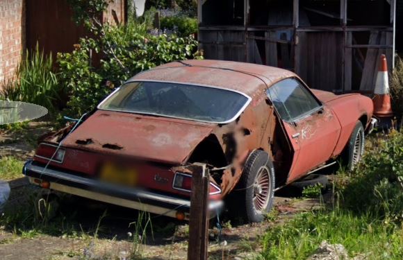 Classics left to die/rotting pics - Vol 2 - Page 339 - Classic Cars and Yesterday's Heroes - PistonHeads UK - This is a color photograph featuring an old, abandoned car parked in what appears to be a rural setting. The car is red and rusted, indicating its age and disuse. Its bodywork shows signs of significant corrosion, with rust spreading across various parts. In the background, there's a small building and a patch of greenery. There's also a vintage truck visible in the far distance. The overall atmosphere of the image suggests a scene of abandonment or neglect.