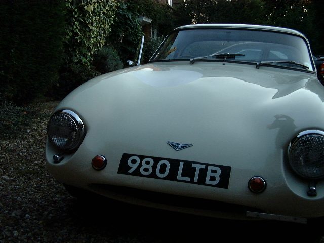 Early TVR Pictures - Page 72 - Classics - PistonHeads - This image presents a vintage car parked on a gravel driveway. The car, which has a reflective finish on its white body, has a black and silver number plate reading "980 LTB". It features two large round headlights at the front, a silver emblem on the hood, and a compact trunk in the back. The vehicle appears to be a convertible, and there's an object with a camera-like symbol in the foreground. The background is characterized by dark and light green hedges, which partially obscure the view of a building. The overall setting indicates a suburban residential area.