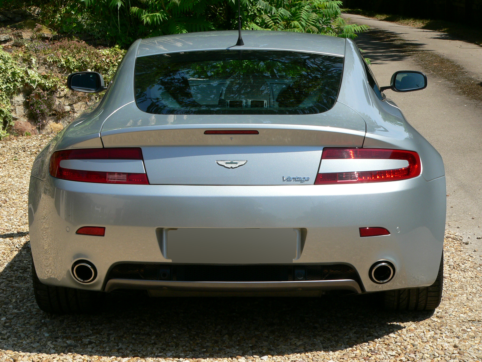 Where are the Titanium Silver Cars (Vantages) ? - Page 2 - Aston Martin - PistonHeads - The image shows a close-up of the rear view of a silver four-door sedan, featuring the tail lights and bumper, parked on a gravel surface next to a paved road. The surroundings are natural, with a roadside banking and a thicket of plants and bushes. There is a clear sky suggesting it's daytime. There are no texts visible in the image. The lighting and focus are tight, emphasizing the shiny metal and the reflective surface of the car.