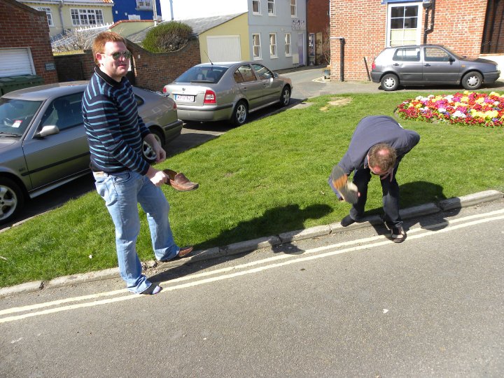 Funniest Pistonheads Quote - This image features a street scene where two men, possibly friends, are engaged in a playful interaction. The man on the left, with his foot up against the curb, appears to be throwing shoes toward the other man, who is slightly bent. They are standing on a sidewalk, framed by parked cars. In the background, there's a brick building, adding an urban touch to the scene. Flower planters can be seen as well, suggesting a well-maintained neighborhood. Despite the casual activity, the scene has a relaxed and friendly vibe.