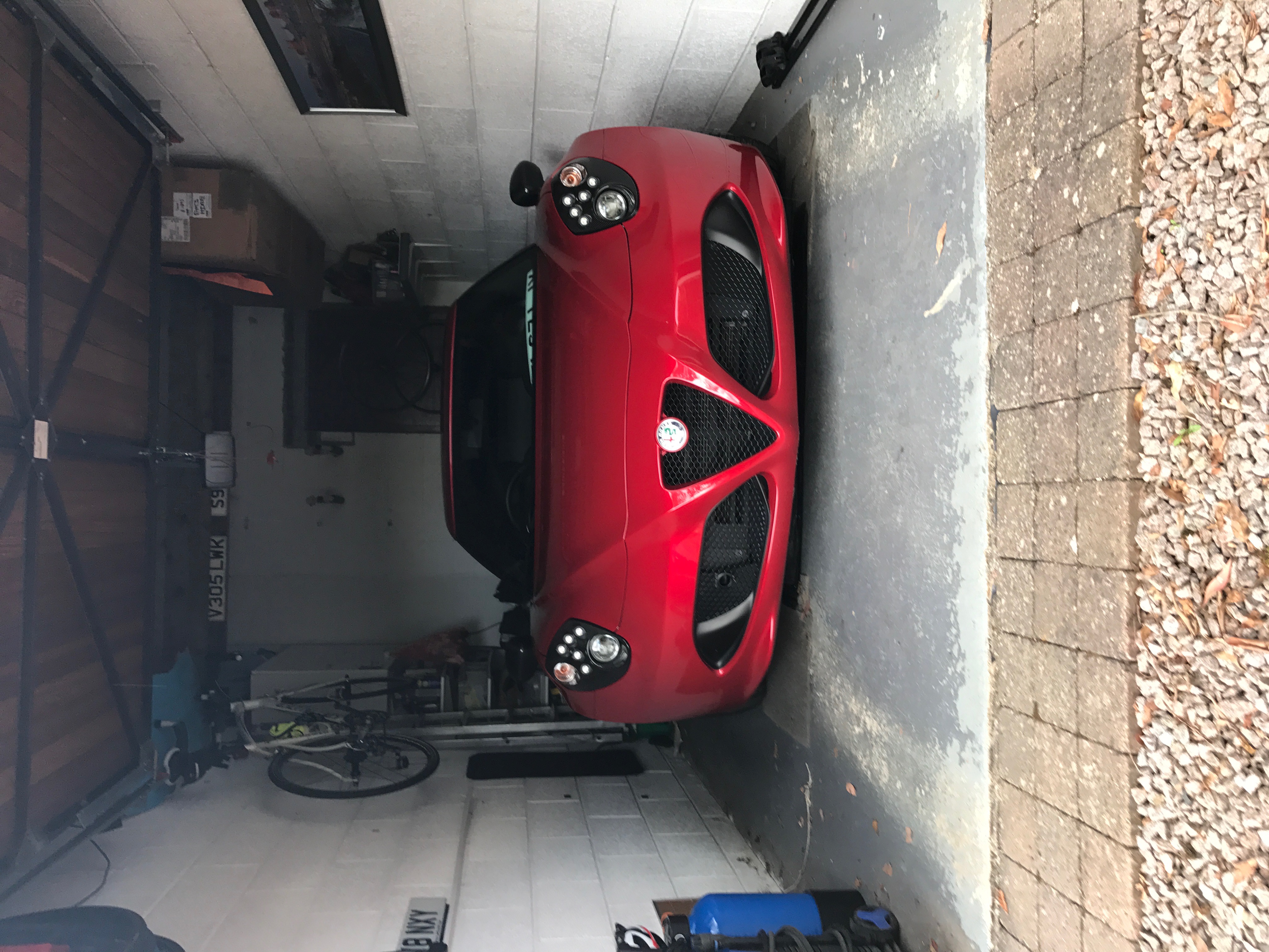 Let's see your Alfa Romeos! - Page 107 - Alfa Romeo, Fiat & Lancia - PistonHeads - The image depicts a vibrant red sports car that appears to be parked in a garage with a brick floor. The car is situated in a space that also houses a bicycle, indicating a multi-purpose use for the garage. In addition, there are various items scattered around the garage, including what seems to be bicycle parts and a piece of equipment or machinery. The car itself showcases a sleek design with a sloping roofline that is characteristic of sports cars, although the specific make and model are not identifiable. The garage, with its brick floor and array of objects, provides a tangible contrast to the smooth and polished look of the red sports car.