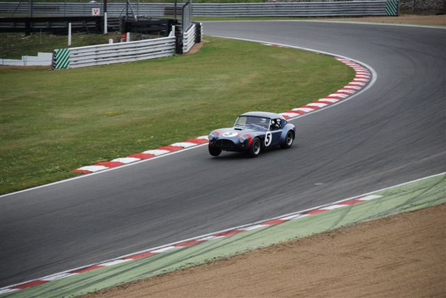 Masters Hatch Brands Festival Pistonheads - The image captures an intense moment at a car race. The main focus is a vintage race car, painted in a striking combination of red and white, positioned in the middle of the track, appearing to exit a bend. The car is speeding through a tight turn, its left side leaning widely into the turn, displaying the dynamic forces at play. The background reveals a bustling pit lane, filled with various support vehicles, adding to the atmosphere of a high-stakes competition. The overall composition of the image conveys the exhilarating experience of a race event.