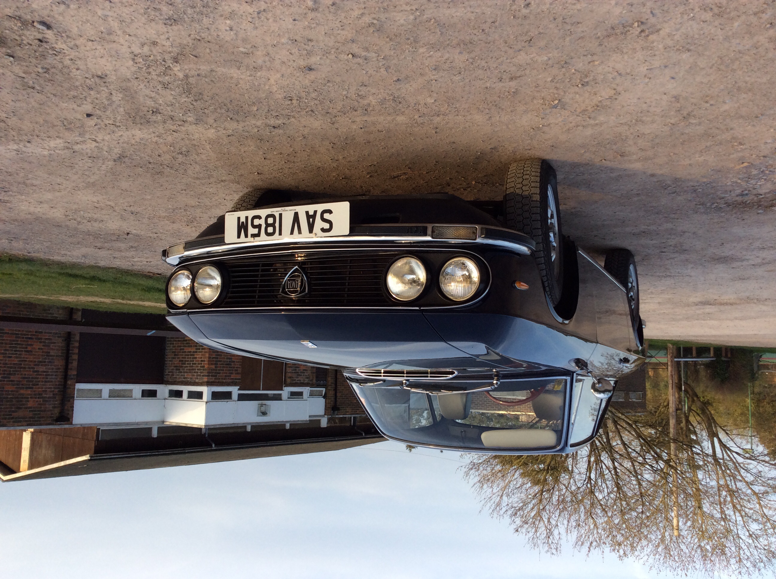 Lets see your Lancia's! - Page 23 - Alfa Romeo, Fiat & Lancia - PistonHeads - The image captures the top-down view of a classic car. The car is parked on what appears to be a lot with sparse vegetation in the background. The contrast between the sky and the ground is evident. The black and white license plate on the car stands out against the blue of its body. The reflection of the sky in the windows of the car adds to the composition of the photograph.