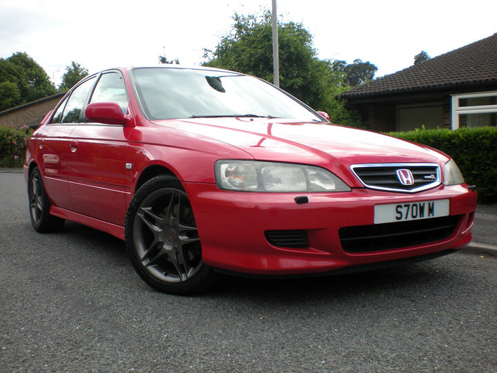 Get your car rated! - Page 54 - Readers' Cars - PistonHeads - The image depicts a vibrant red Honda Accord car parked outdoors. The car, a two-door version, is positioned on a tarmac driveway. Prominently displayed are a metallic grille emblazoned with the Honda logo and distinct headlights. The vehicle features a set of five-spoke alloy wheels. There is a residential building, possibly a house or a structure, partially visible on the left side of the image. The scene captures a sunny day, with daylight illuminating the car and highlighting the car's details.