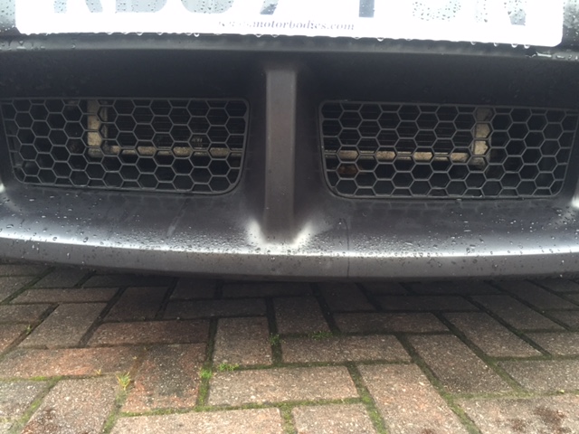 Older charger not intercooled - Page 1 - HSV & Monaro - PistonHeads - The image shows a close-up of a car's front grille, which features a large mesh design, possibly where the license plate is mounted. The image is taken from a low angle, highlighting the grille and car's bumper. It appears to be a rainy day as the grille and bumper are wet, suggesting the car has recently driven through some moisture or rain has just fallen. There is a distinctive arched shape to the grille, which adds to the close-up composition. The colors in the image are relatively muted, with the car's black and gray color scheme being the most prominent.