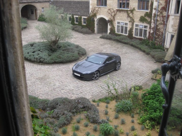 Manor Pistonheads Whatley - The image presents a tranquil street scene viewed from a window. A sleek black sports car is parked on the neatly arranged cobblestone driveway, positioned in a circular motion orderly enough for a level playing field. The car is next to a large gray building, possibly a mansion, characterized by classical architectural elements on brick pillars. The front yard is adorned with a variety of small, green shrubs, contributing to the overall calm and well-maintained aesthetic of the residential area.