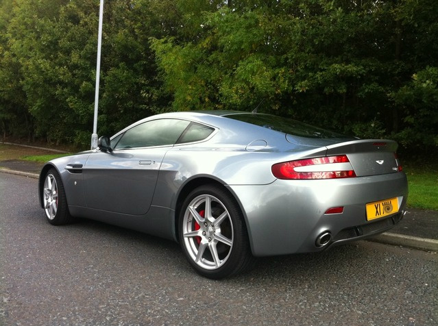 New Vantage owner. - Page 1 - Aston Martin - PistonHeads - The image presents a stylish, metallic gray luxury car parked on the side of a roadway. The car's sleek lines and high gloss paint catches the sunlight, highlighting its shiny surface and reflecting some of the surrounding greenery. The vehicle has a distinctive rear wing and a license plate. It is situated under a streetlight, suggesting it could be in a driveway or a shaded parking area. The license plate reads "XII A15," indicating it might be a model from the U.K. The car's position on the street, its design, and the surrounding trees create a serene and elegant atmosphere.