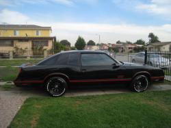 Pistonheads Speed Obscure - The image depicts a black, two-door car parked on a driveway. The driveway is adjacent to a lawn with grazing patches of grass. In the background, there are residential houses and an iron gate. The sky above is partly cloudy, suggesting a calm day. The car appears to be in good condition, with shiny surfaces indicating regular maintenance.