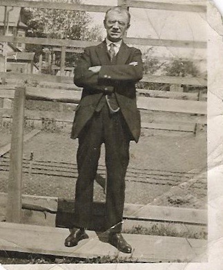 The image is a black and white photograph featuring a man dressed in formal attire, standing confidently with his arms crossed. He wears a suit with a tie, indicating a sense of formality or professionalism. The background of the photo suggests an outdoor setting, possibly a ranch or farm, with wooden fencing and a dirt surface. The photo appears to be older, as indicated by the grainy texture and monochrome scale of the image.