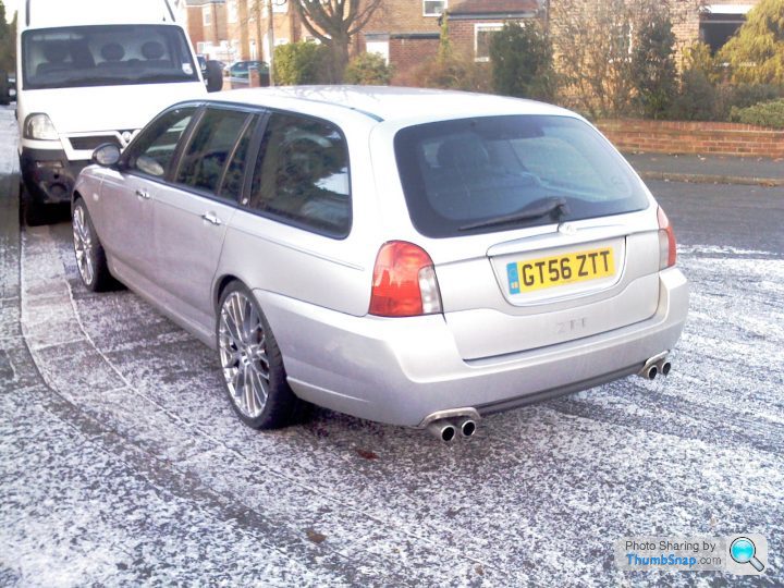 Service Pistonheads Costs - The image depicts a silver car parked on a snowy street. The car is positioned behind another vehicle, its rear facing the viewer. The vehicle's license plate reads "GT56 ZZT". The snow appears freshly fallen, indicating cold weather conditions. In the background, there's a white truck visible. The setting suggests a residential area, possibly a neighborhood street, given the presence of parked vehicles and a glimpse of houses.