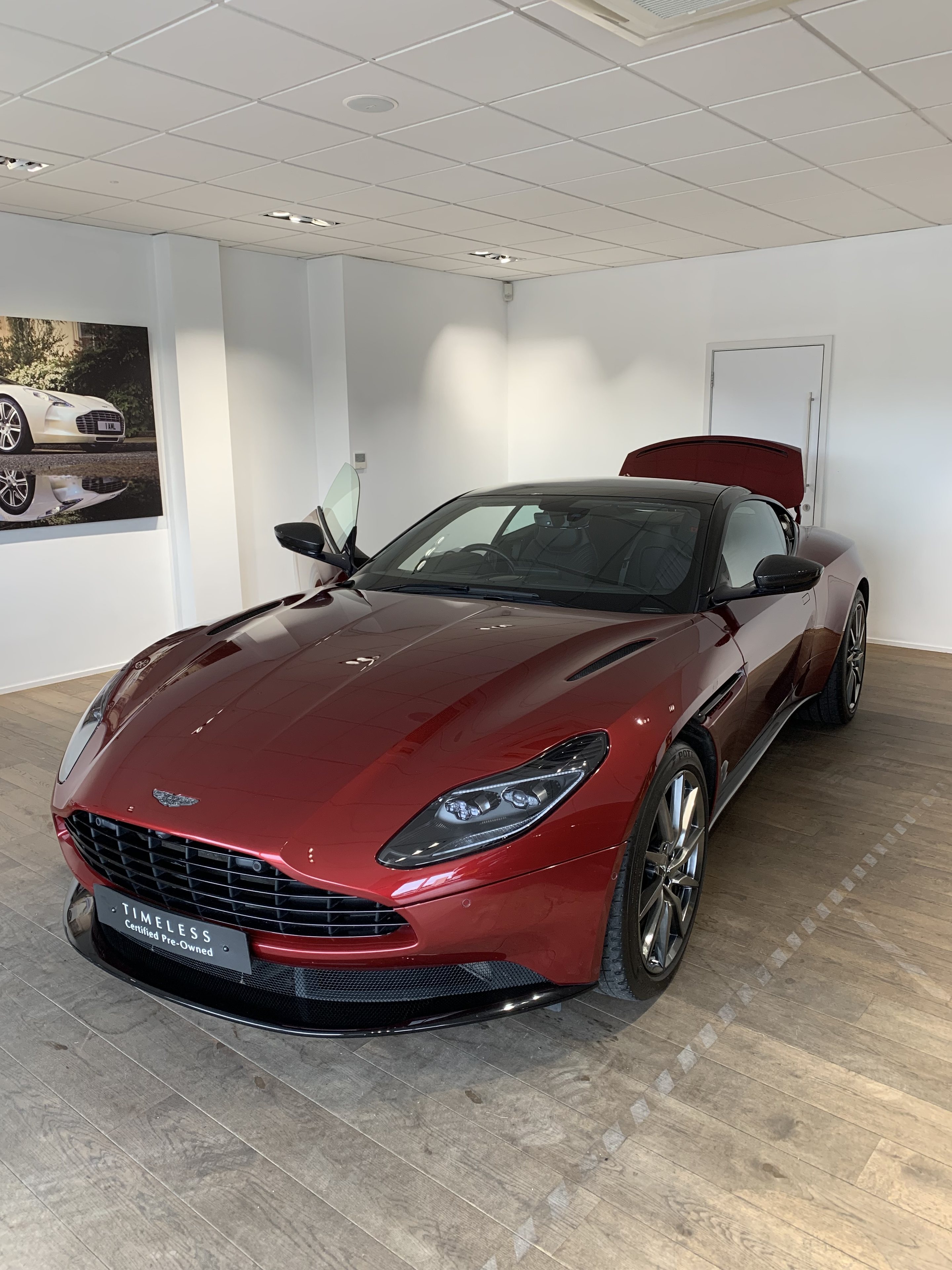 F90 M5 or DB11? Used purchase quandary... - Page 6 - Car Buying - PistonHeads UK - This image depicts a showroom or sales area where an Aston Martin car is on display. The car, which is the focal point of the image, has an open hood, revealing its engine, and is positioned as if ready to be driven. The vehicle is sleek and appears to be a high-end model with a distinctive design typical of luxury sports cars. The interior of the showroom is well-lit and minimalistic, allowing the car's features to stand out prominently. There are no visible texts or distinctive brands other than the Aston Martin logo on the vehicle.