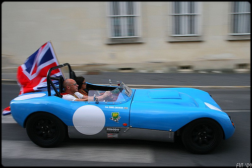 Pistonheads - This image captures a vibrant scene on a city street. At the center of the frame is a convertible sports car painted in a striking shade of blue. The car is driven by a man in a red shirt, who is joined by a woman seated in the passenger seat. Adding a patriotic touch to the scene, a flag featuring red, white, and blue is proudly displayed on the back of the car. The car is in motion, driving past the rear view, creating a blurring effect that enhances the sense of speed. The overall image conveys a sense of thrill and adventure.