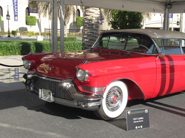 Classic Pistonheads Emaar - The image displays a classic, vintage car on display, viewed from a three-quarter angle. The car is positioned under a stone archway. The vehicle has a glossy red exterior and white-walled tires, and is further accented by its chrome details, adding to its antique appeal. To the left, part of a green lawn is visible, and the ground surrounding the car appears to be a dark surface. There's a plaque in front of the car on the ground, but the text on the plaque is not legible in the image. The car's model is not identifiable from this angle and perspective.