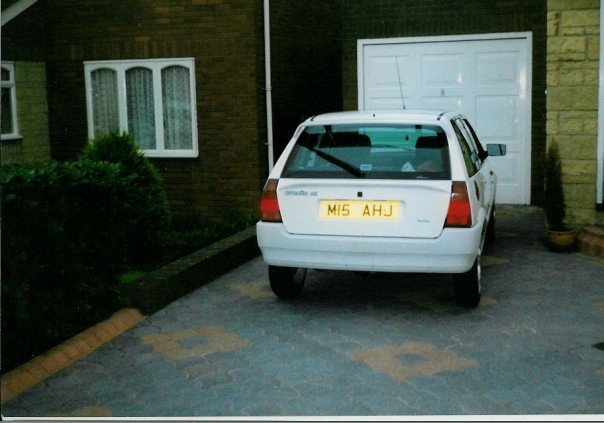 My Car - Page 1 - Readers' Cars - PistonHeads - The image shows a white car parked on a paved driveway of a two-story brick house. The bricks have a red-brown color. To the left of the driveway is a small, green bush. A white garage door is open behind the car, allowing a view into the interior of the garage. Another set of windows is visible, situated on the brick house. The car has a yellow license plate with the text "MS AHJ," which could indicate a registered vehicle in a specific jurisdiction.