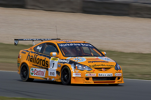 Pistonheads - The image displays a bright yellow race car on a fast-paced track. The car features a number at the front and a variety of logos and decals adorning its orange body. There is a driver within the cockpit, but the driver is not facing forward which indicates this could be a still image, possibly from a race or a promotional poster. The track appears smooth and well-maintained, with a blur effect indicating high speed.