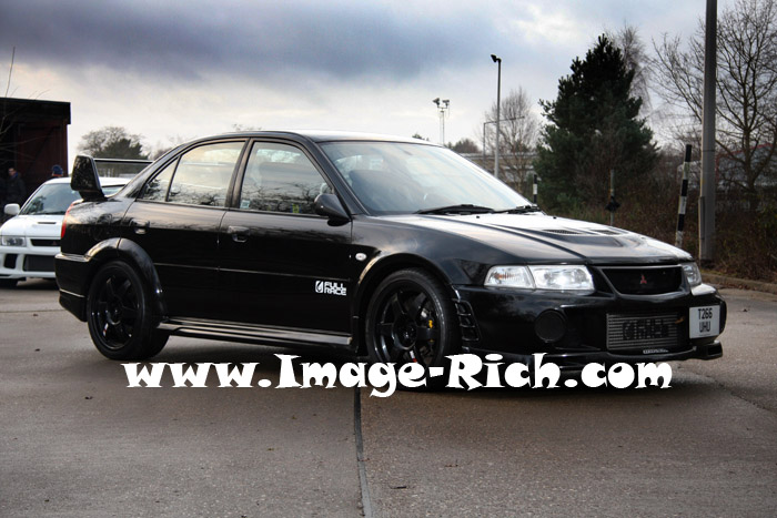 Newley Registeredpics Pistonheads Evo - The image features a black car with visible modifications, parked on an affordable auto insurance website. The car appears to be a compact, possibly a hatchback or coupe, with a unique rear bumper, side skirts, and a lowered stance. There are several cars parked in the background with people standing nearby, indicating this might be a public parking lot. The sky in the background suggests it could be an overcast day.