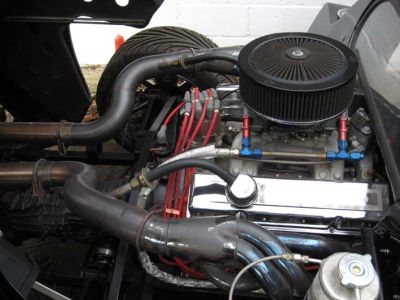 Help identifying engine - 440 Magnum? - Page 1 - Yank Motors - PistonHeads - The image features an engine compartment of a car, focusing on the mechanical parts. A large black fan is prominently visible at the top right with a red and black fan wheel. Adjacent to this is a red wire connected to the fan. Further down, there's a coil of copper tubing leading to a metallic radiator. There are blue hoses connecting the engine to the radiator, and a silver metal hydraulic accumulator is attached to the right. The background is a plain white wall, suggesting an indoor setting, likely a garage or workshop.