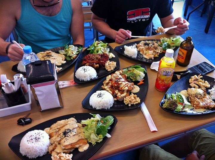 A group of people sitting around a table eating food - Pistonheads - In the image, a lively dining scene is depicted. Three individuals are seated around a dining table, engrossed in their meal. They are served a variety of Asian cuisine dishes. In particular, the dishes include Chinese-style shrimp, accompanied by steamed white rice. The table is adorned with an array of condiments and drinks, with bottles and cups scattered across the table. The atmosphere appears casual and relaxed, with one person's mobile phone placed on the table.