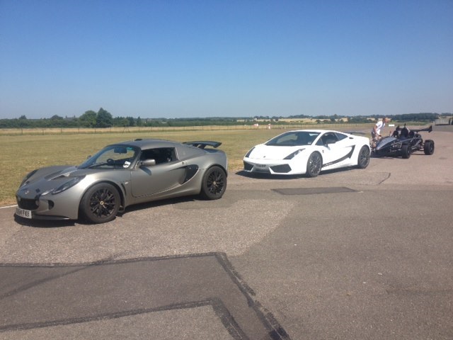 Track Day options. - Page 1 - Kent & Essex - PistonHeads - The image captures a scene at a race circuit, where three high-performance sports cars are parked. A silver Audi R8 is sleek and streamlined, exuding a sense of speed and power. Beside it, a pristine white Ferrari stands in stark contrast with its polished exterior. Not far from them, a black Lotus is parked, its aerodynamic design hinting at its high-speed capabilities. The setting offers a clear view of the circuit ahead, with a beautiful expanse of grass forming the backdrop.