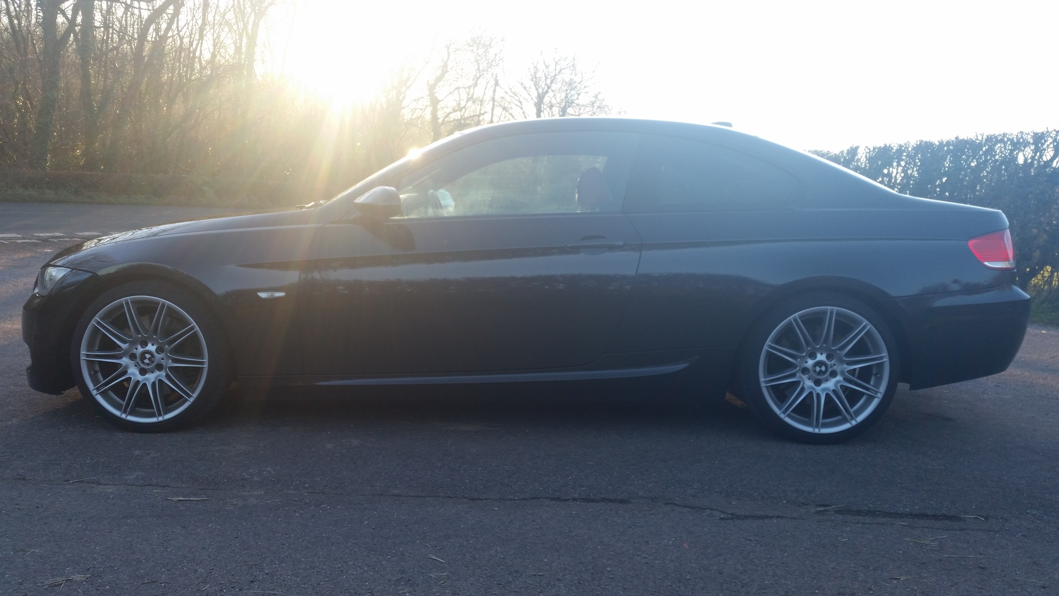 Show Me Your BMW!!!!!!!!! - Page 299 - BMW General - PistonHeads - The image shows a dark-colored sports car on a paved road during the sunset. The car is positioned at an angle, highlighting its profile and the BMW emblem on the trunk. The surrounding environment includes a line of trees and a clear sky. The sunlight shines through the rear passenger side window and onto the car, creating a lens flare effect. The setting appears peaceful with a blend of natural light and the car's silhouette.